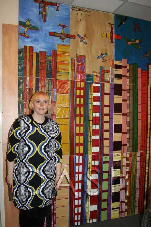 Barriers Artist, Rowena Keaveny stands next to her collaborative painting.  The painting consists of nine separate canvases, which make a block - 3 tall and 3 high.  The painting uses all colours and shows tower blocks on the bottom two rows, on the top row is the top of the tower block and in the sky above are airplanes.  The canvases were all painted by artists involved in the Barriers project.  Photograph by Paula Dower copyright DASh.